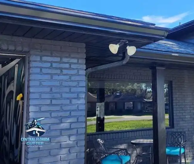 "Black seamless aluminum gutter and downspout installed on a grey brick home with matching dark soffits and outdoor lighting."