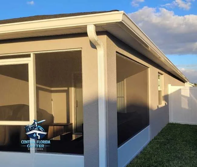 Corner view of a screened-in patio on a gray house featuring newly installed white gutters and a matching vertical downspout.