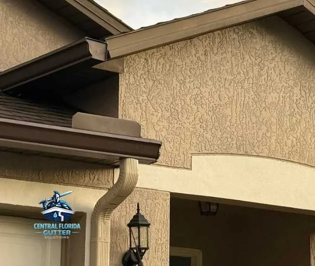 Upward view of dark brown gutters and a matching tan downspout installed on a stucco home with a dark shingle roof and a classic outdoor wall lantern.