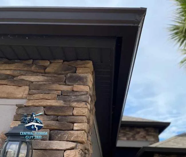 Upward view of a dark brown gutter and matching soffit system installed on a home with a stone-cladded exterior wall and an outdoor light fixture.