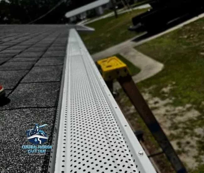 Long-perspective view of white perforated gutter guards installed along the edge of a gray shingle roof, with a ladder and residential lawn in the background.