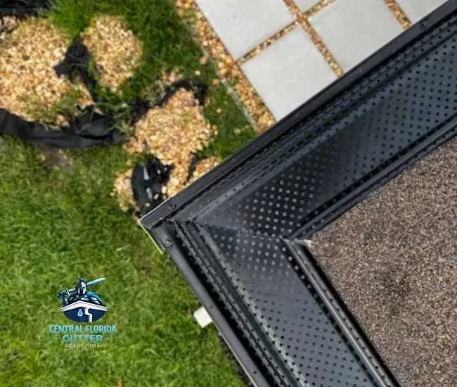 Top-down view of a roof corner featuring black perforated gutter guards installed over a dark shingle roof, overlooking a lawn with gravel patches.
