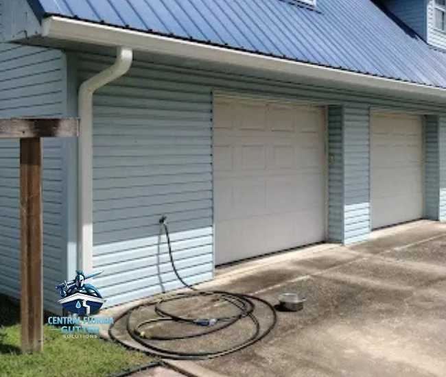 A light blue house with a blue metal roof featuring white gutters and a downspout next to a white two-car garage.