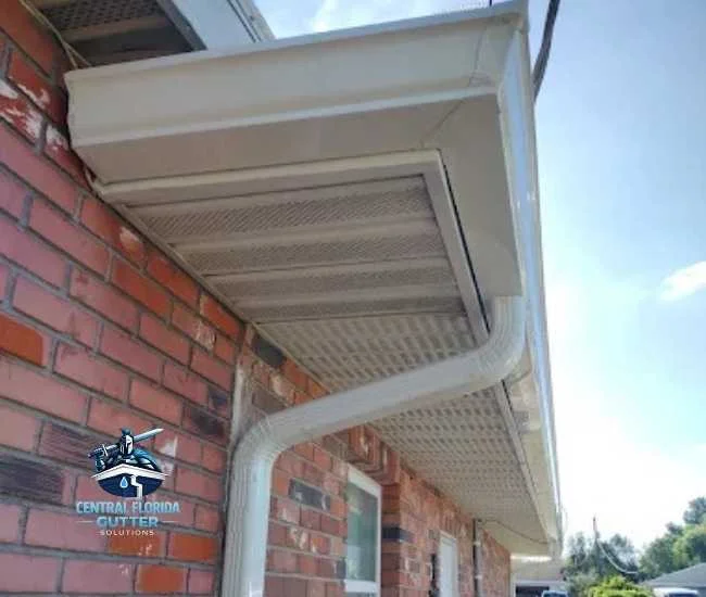 Low-angle shot of a brick house featuring newly installed white gutters and a curved downspout against a light gray soffit.