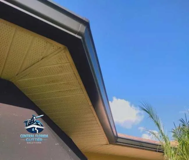 Upward view of dark gutters and tan perforated soffit panels installed on a home under a clear blue sky.