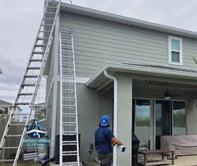A worker standing next to tall extension ladders leaning against a two-story gray house while performing gutter maintenance.