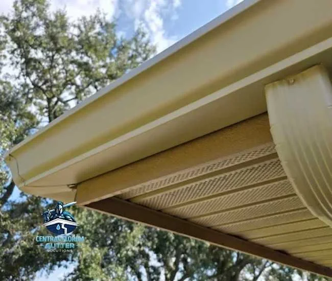 Upward view of beige gutters and matching perforated soffit panels on a home with lush green trees in the background.