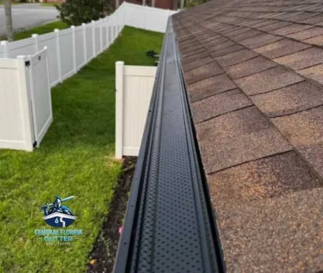 Top-down view of black perforated gutter guards installed along the edge of a brown shingle roof, overlooking a green lawn with a white vinyl fence.