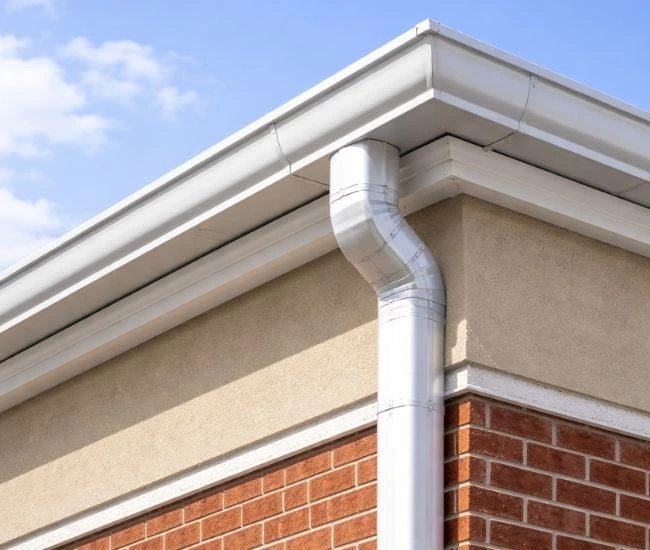 A low-angle shot of a commercial building corner featuring a sleek, metallic gutter and downspout system. The building features a brick facade topped with a white soffit and fascia. The gutter runs along the roofline, transitioning into a cylindrical downspout that extends down the brick wall. The scene is set against a clear, vibrant blue sky